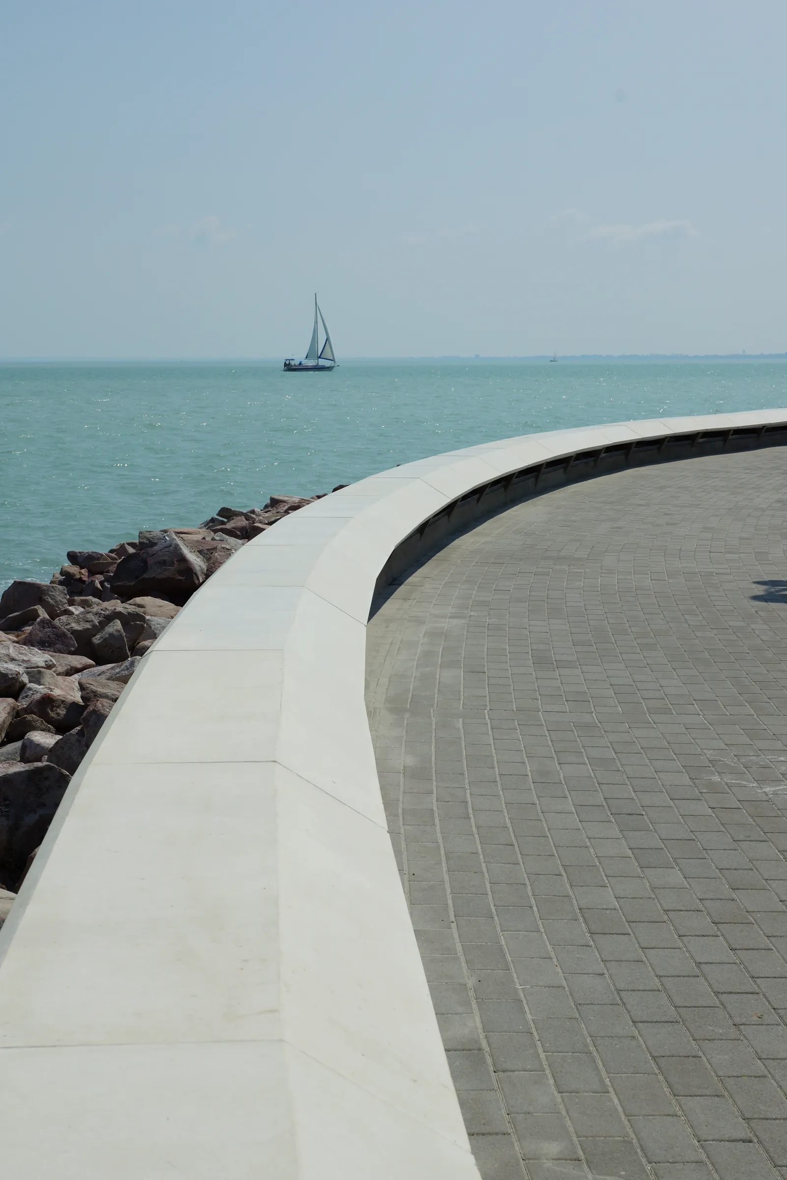 The curved cast-stone parapet wall and paving of the pier head, with a sailboat on Lake Balaton beyond the rip-rap shoreline