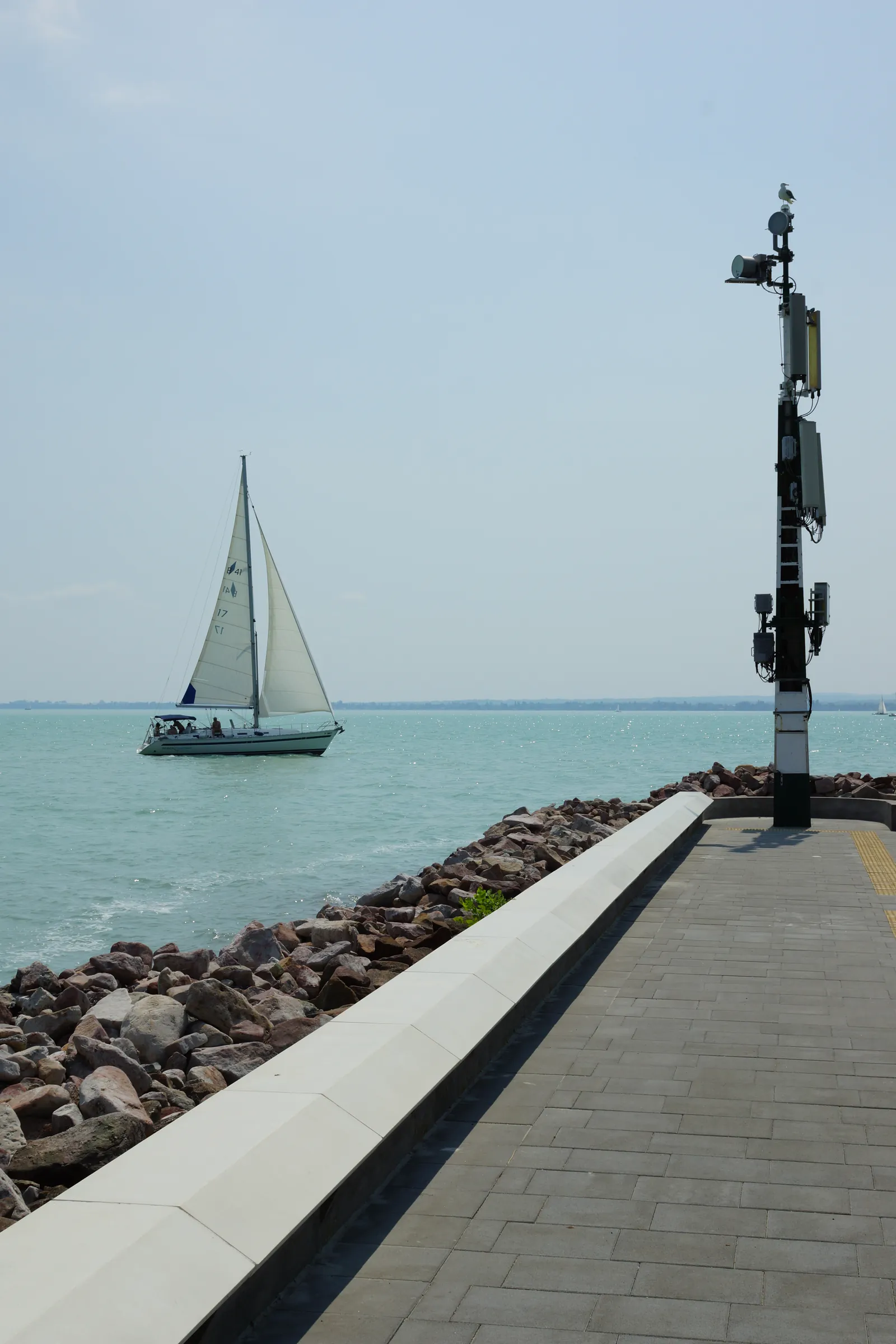 The port's pier head with its cast-stone edge facing the water and a navigation mast, a sailboat in the background