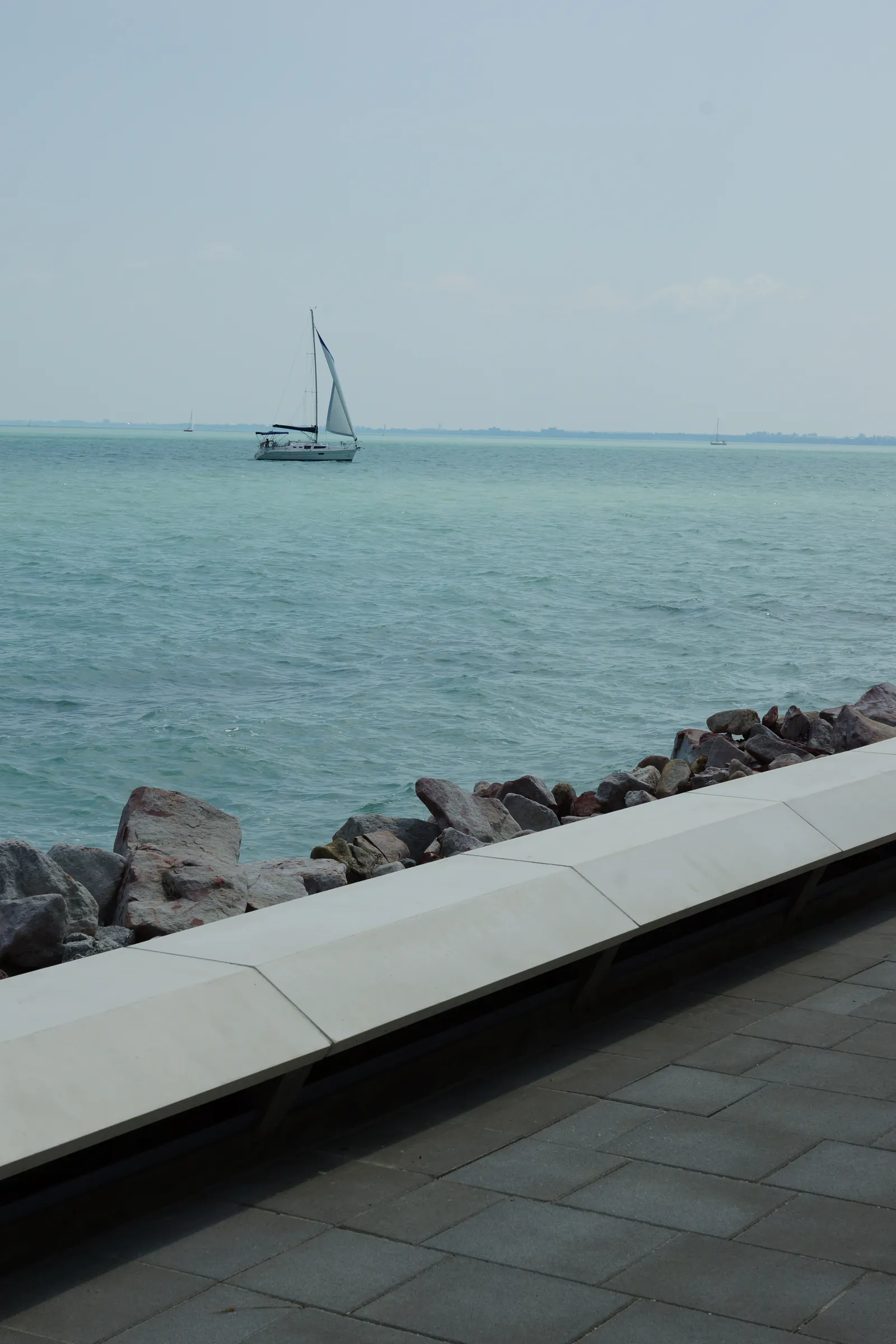 The low cast-stone parapet wall and paving of the pier head by the rip-rap shoreline, with a sailboat on Lake Balaton