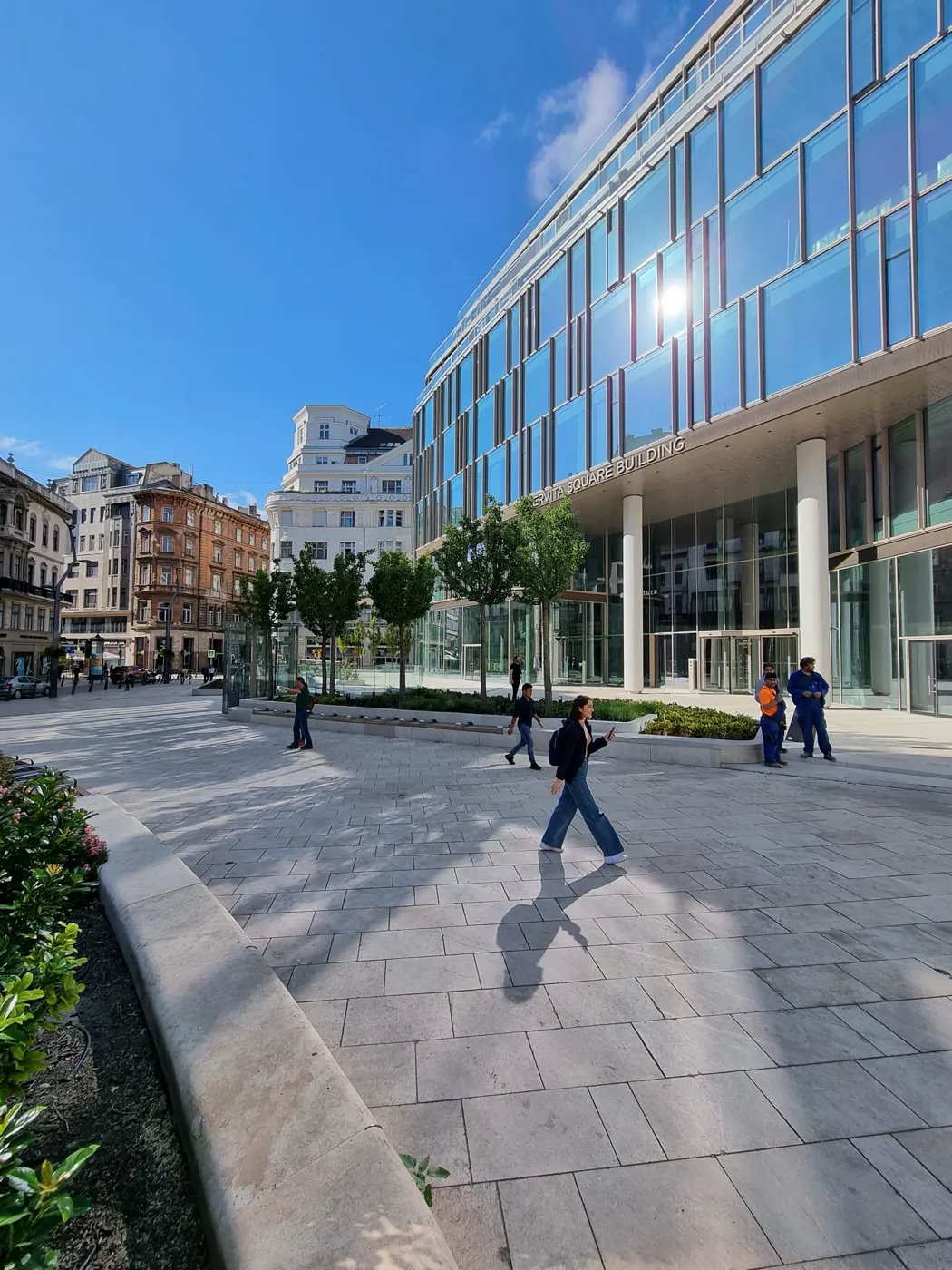Gyalogosok a kiemelt fogadótéren, a növénykazetta íves pereme és a Szervita Square üveghomlokzata