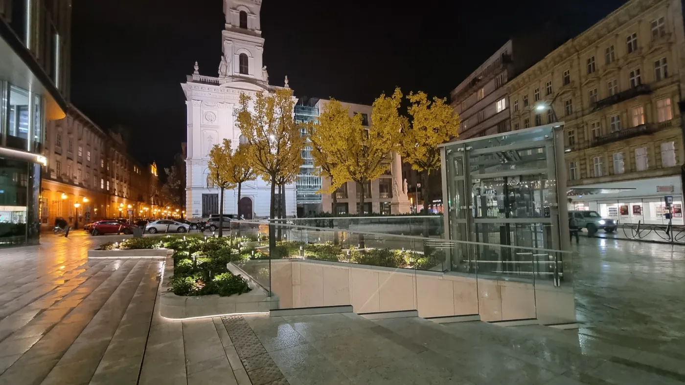 A fogadótér este: megvilágított növénykazetták, őszi sárga páfrányfenyők, üveg liftház és a Szervita templom